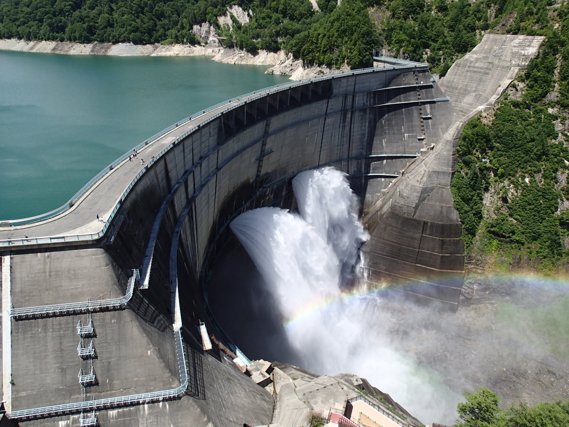 Kurobe Dam mid water-discharge, a mist plume rising over the arch dam with green slopes around it